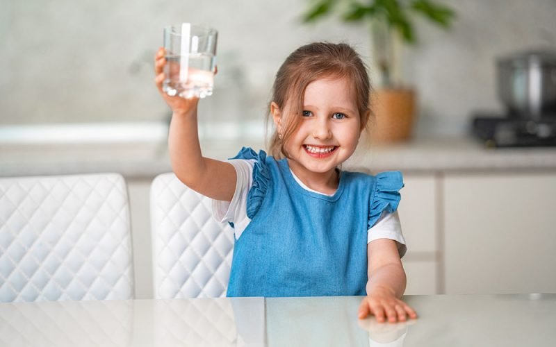 Cute little girl drinking water in the kitchen at home. The little girl is smiling happily, holding a glass of clean water and wants to drink it. Water balance. Prevention of dehydration